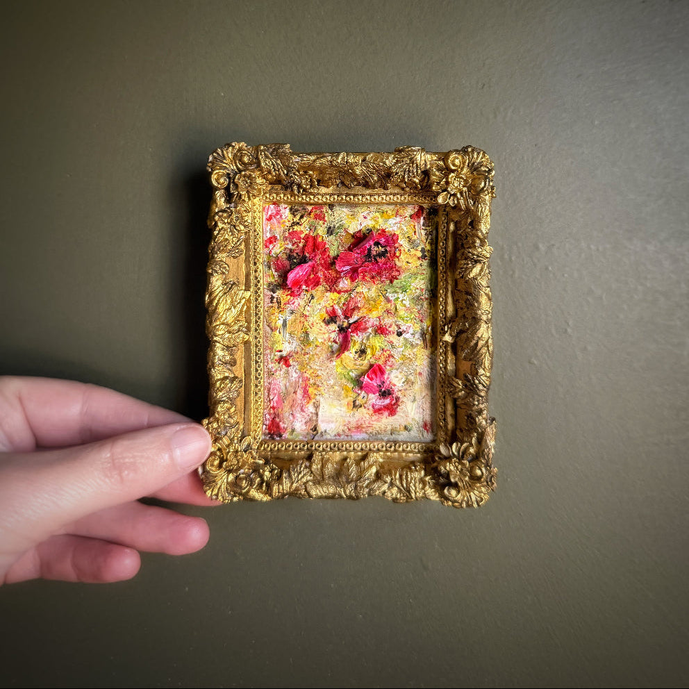 Hand holding a small floral painting in a gold frame against a plain background