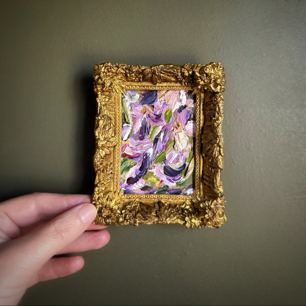 Hand holding a small framed painting with a gold frame against a plain background