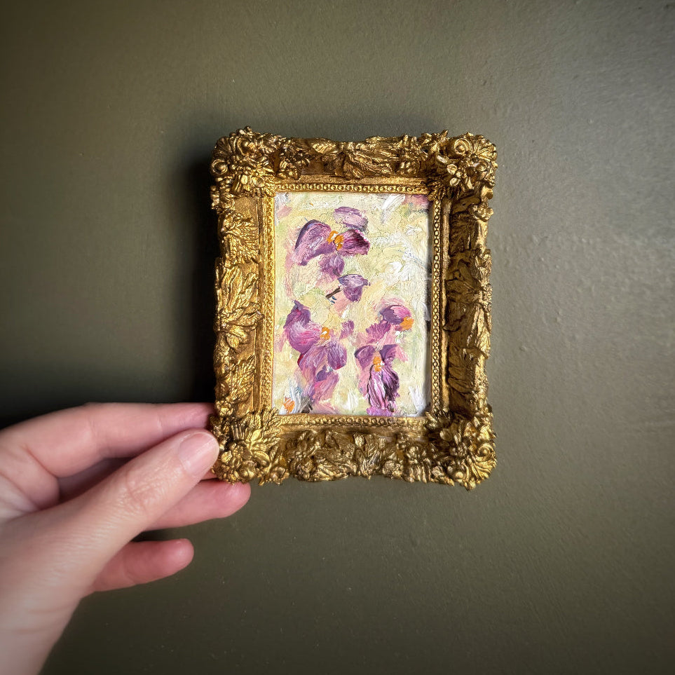 Small painting of purple flowers in a gold frame held against a dark background
