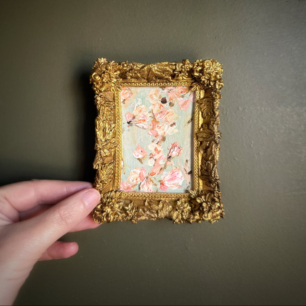 Hand holding a small ornate gold frame with a floral painting against a dark background