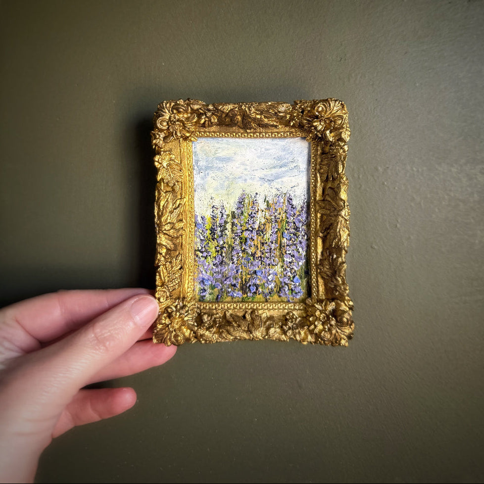 Hand holding a small gold-framed painting against a plain background