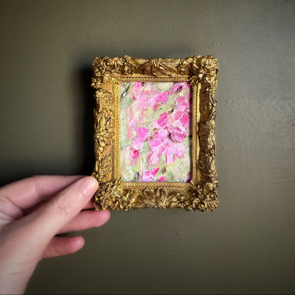 Hand holding a small gold-framed artwork with pink floral design against a dark background