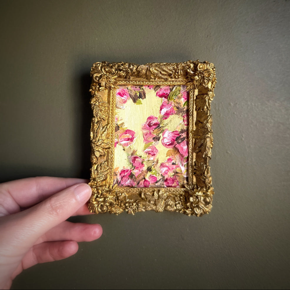 Gold-framed artwork with pink floral design held by a hand against a dark background