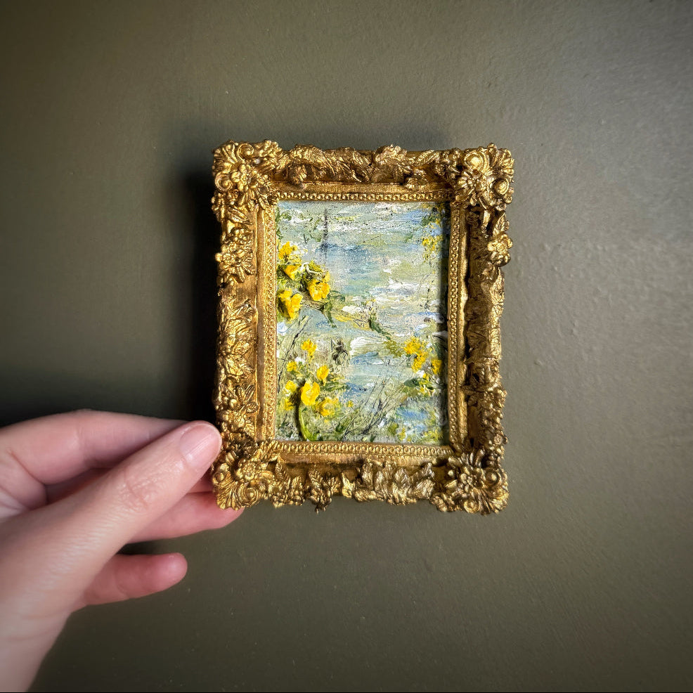 Small oil painting in a gold frame held by a hand against a dark background
