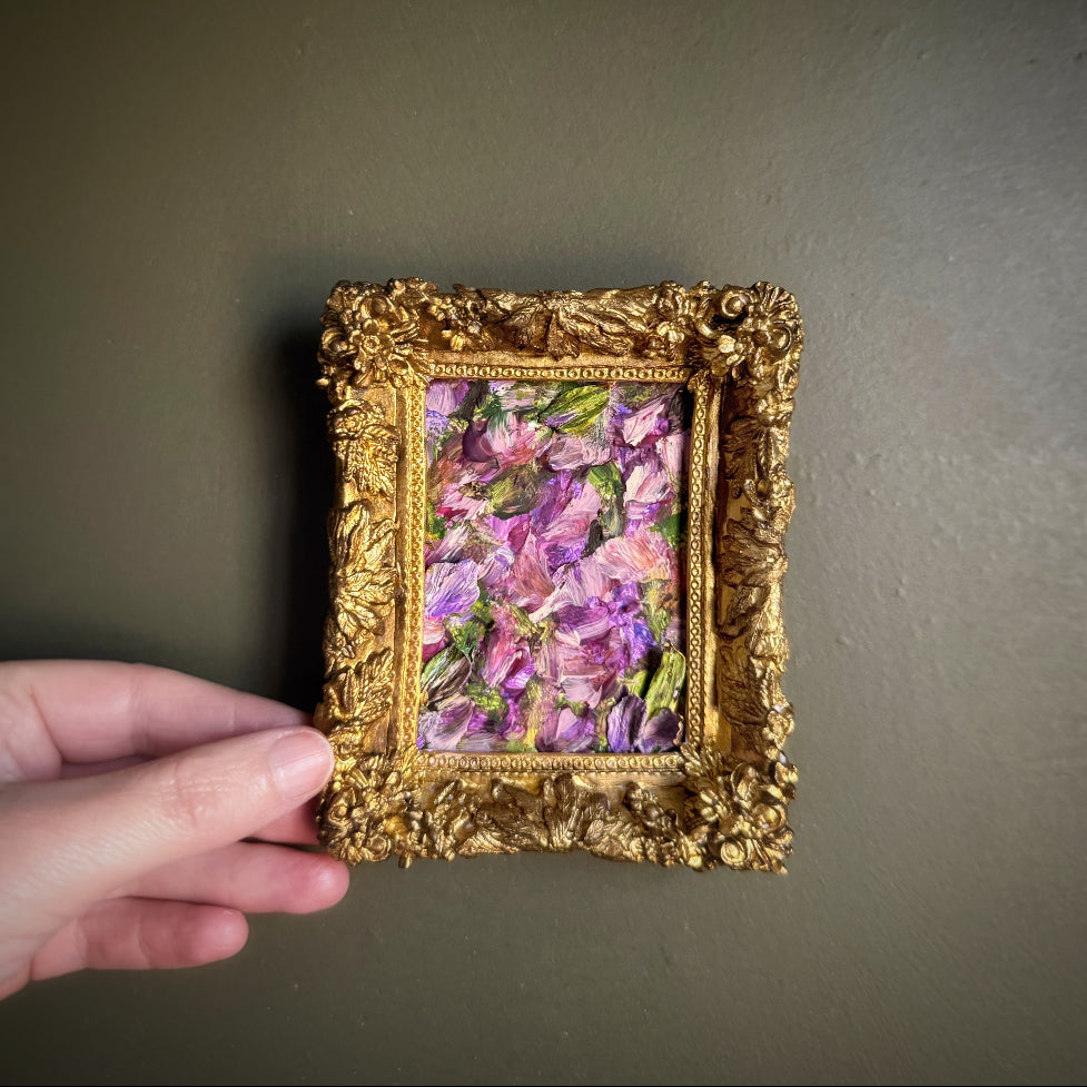 Hand holding a small floral painting in an ornate gold frame against a plain background