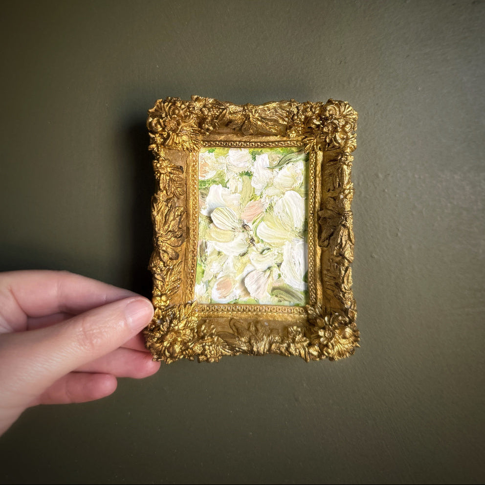 Hand holding a small ornate gold frame with a floral painting against a plain background