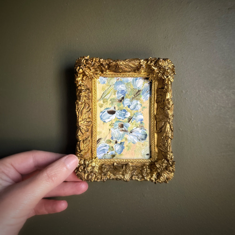 Hand holding a small framed painting with floral design against a plain background