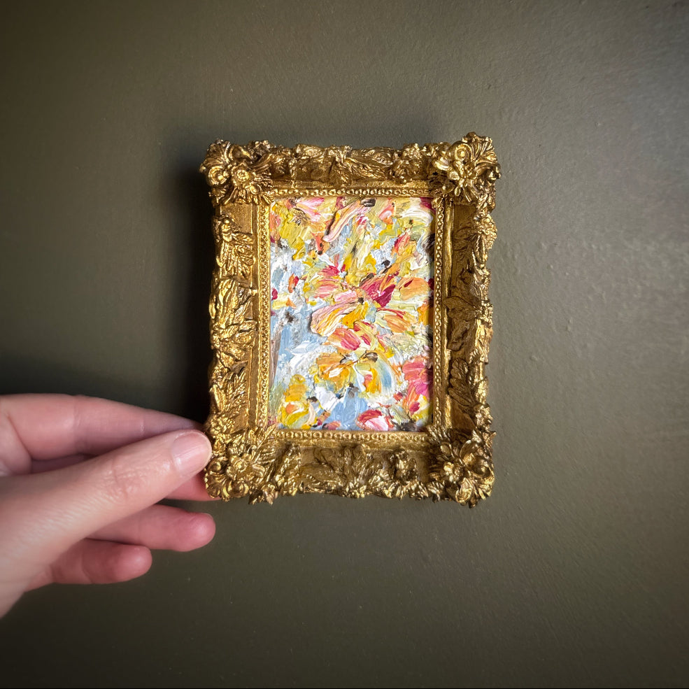 Hand holding a small ornate gold frame with a colorful painting inside against a dark background
